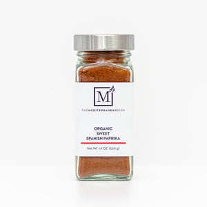 A glass spice jar filled with sweet paprika, featuring a white label with the brand logo 'The Mediterranean Dish' and a silver metal lid, set against a clean white background.