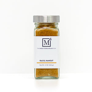 A glass spice jar filled with ras el hanout, featuring a white label with the brand logo 'The Mediterranean Dish' and a silver metal lid, set against a clean white background.