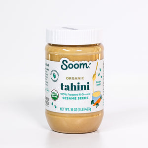 A jar of Soom Organic Tahini Sesame Seeds on a clean white background. The jar contains a smooth, light brown peanut butter-like paste and features a white lid and a white label with green and gold text.
