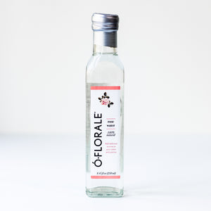 A clear glass bottle of ÉFloraLe Pure Water with a white label featuring pink accents and a silver cap, set against a plain white background.